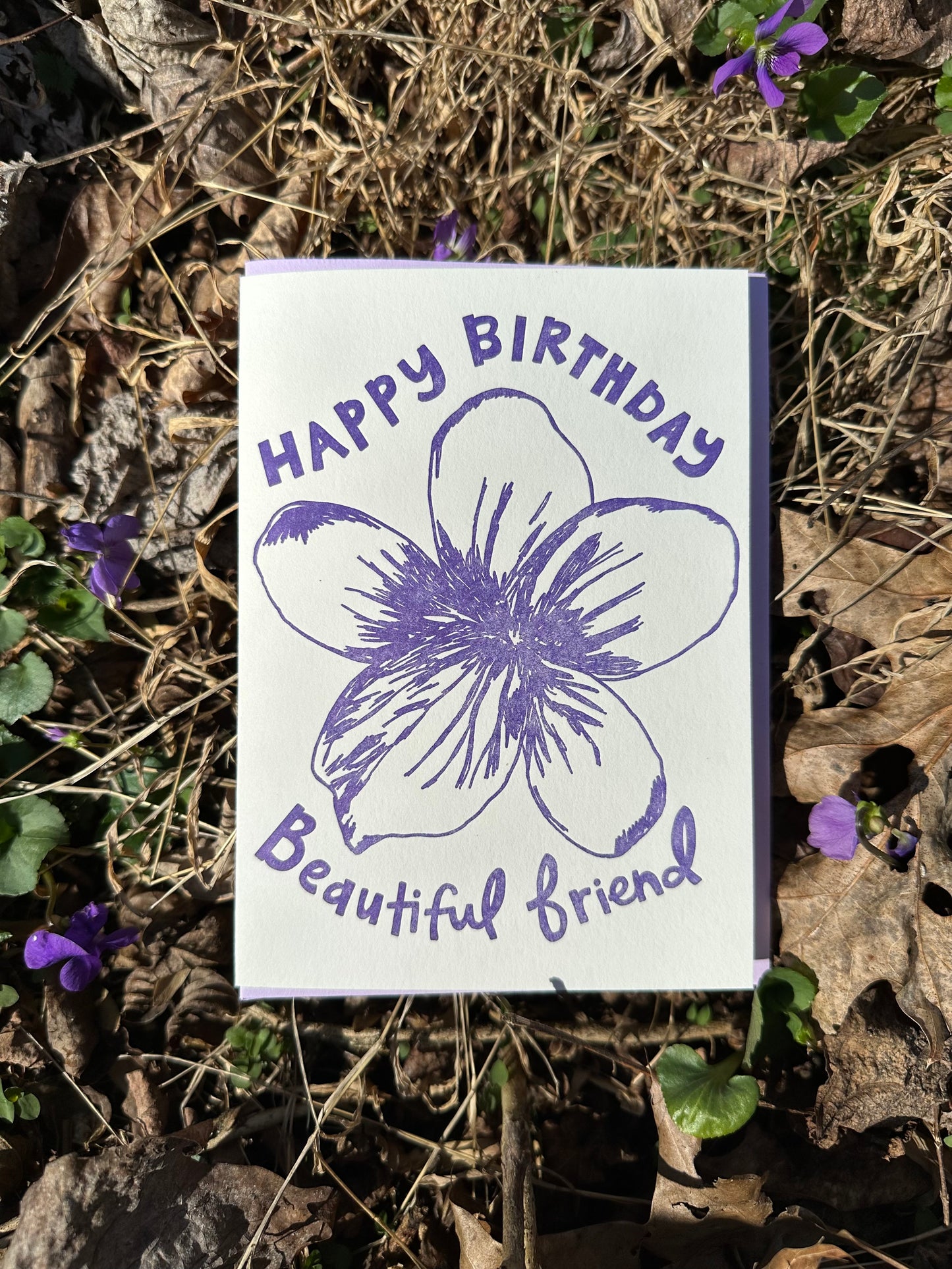 Letterpress greeting card featuring a hand-drawn Violet flower printed in a vibrant purple ink. "Happy Birthday Beautiful Friend!" is written on the top and bottom of the flower in a whimsical hand-drawn text, in the same purple ink. The card is white, blank inside, and is paired with a lilac purple envelope. Card is shown outside on the ground atop some violets on a sunny winter day. 