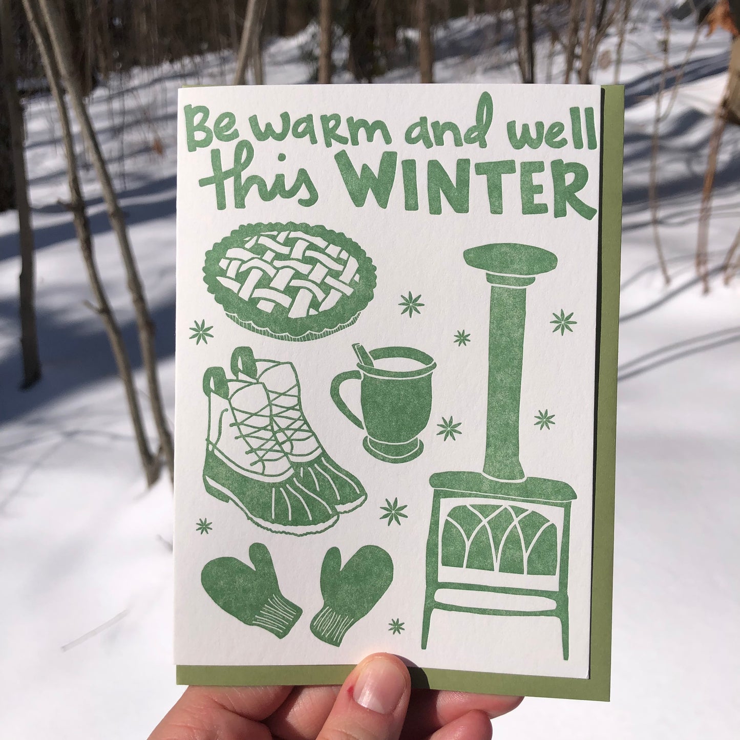 Celebrate Winter with this festive greeting card! This cheerful and cozy card features all the iconic image of the winter season: wood burning stove, star anise, hot cocoa (or cider!), fresh baked pie, mittens, winter boots. Hand-drawn images and typography are letterpress-printed in forest green ink. Green envelope.  Card is shown outside in the winter snow. 