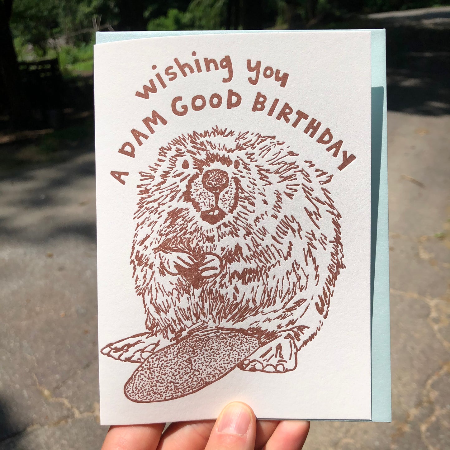 Letterpress greeting card featuring a hand-drawn Beaver printed in an earthy brown ink. "Wishing you a Dam Good Birthday!" is written on the top above the beaver in a whimsical hand-drawn text, in the same brown ink. The card is white, blank inside, and is paired with a light sky blue envelope. Card is being held up outside on a sunny day. 