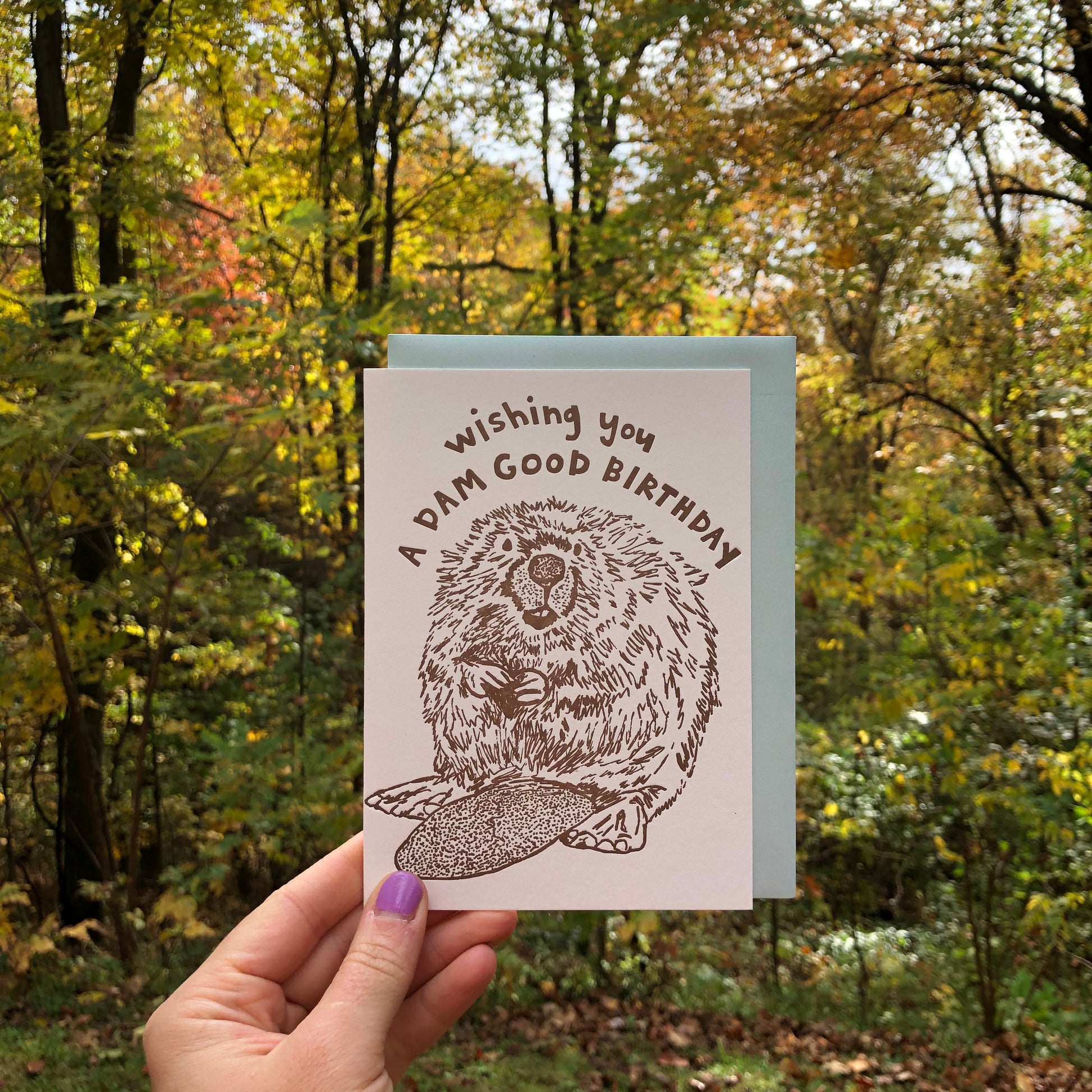 Letterpress greeting card featuring a hand-drawn Beaver printed in an earthy brown ink. "Wishing you a Dam Good Birthday!" is written on the top above the beaver in a whimsical hand-drawn text, in the same brown ink. The card is white, blank inside, and is paired with a light sky blue envelope. Card is being help up in front of an early Autumn deciduous forest. 