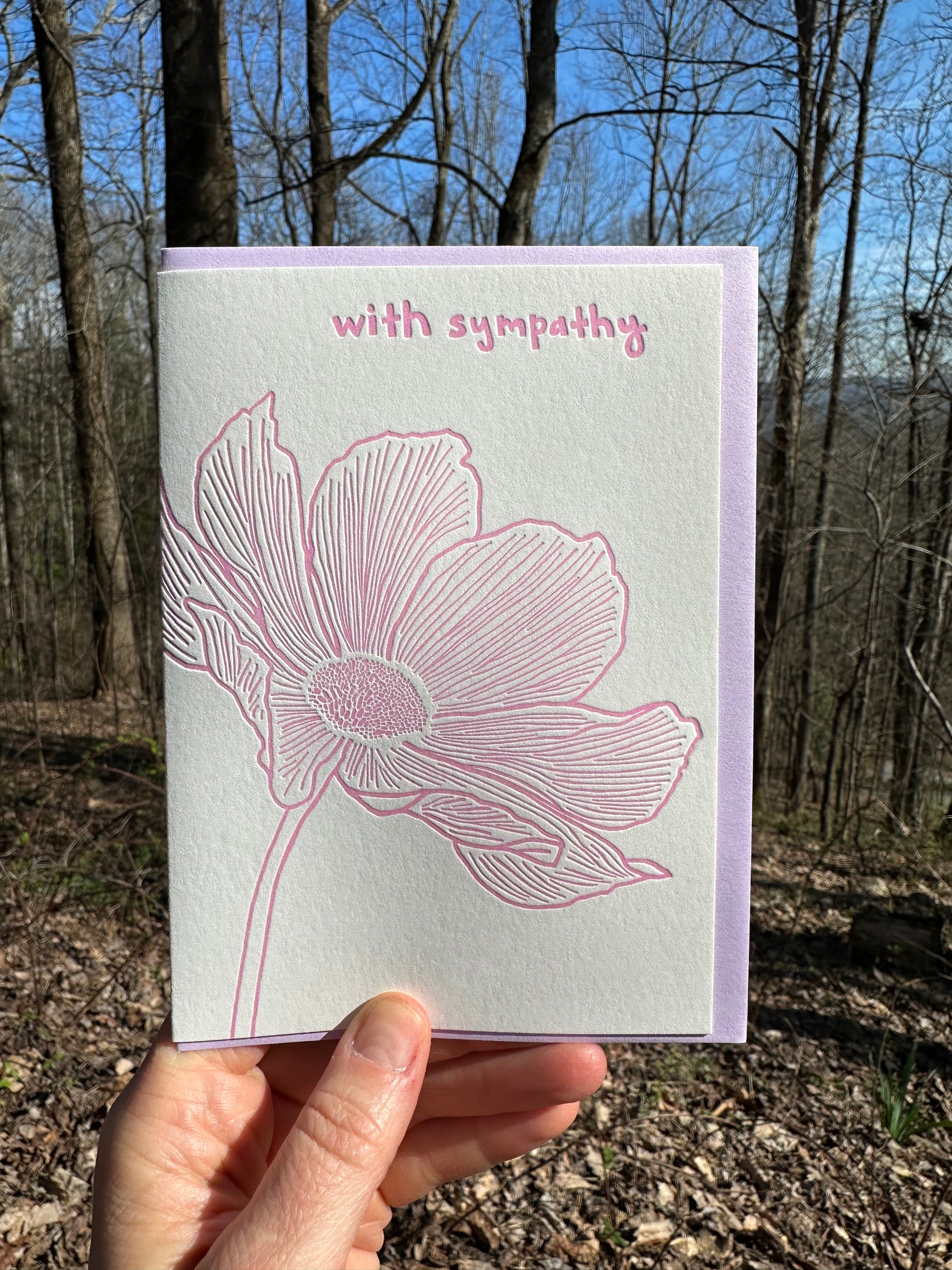 Letterpress greeting card featuring an intricate hand-drawn poppy flower, printed in a vibrant red ink ink. Whimsical hand-drawn text saying "Sincere Condolences" is shown on the right of the card, in the same red ink. The card is white, blank inside, and is paired with a soft yellow envelope. The card is shown outside on a sunny winter day. 