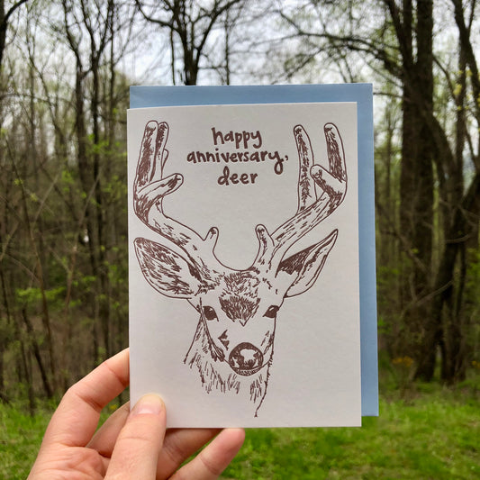 Letterpress greeting card featuring a hand-drawn buck, printed in an earthy brown ink. Whimsical hand-drawn text saying "Happy Annivesary, Deer" is shown inside the deer antlers, in the same brown ink. The card is white, blank inside, and is paired with a light blue envelope. Card is held up outside in front of a spring forest. 