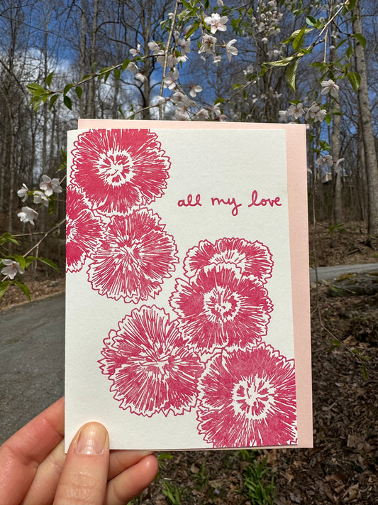 Letterpress greeting card featuring hand-drawn dianthus flowers, printed in a vibrant deep pink ink. The top right of the card says "all my love" in a whimsical hand-drawn text, in the same pink ink. The card is white, blank inside, and is paired with a light pink envelope. Card is held outside by a cherry tree on a sunny day in early spring. 