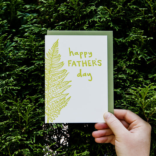 Letterpress greeting card featuring a hand-drawn fern, printed in a vibrant lime green ink. The top right of the card says "Happy Father's Day" in a whimsical hand-drawn text, in the same lime green ink. The card is white, blank inside, and is paired with a green envelope. Card is held in front of a green hedge, zoomed in. 