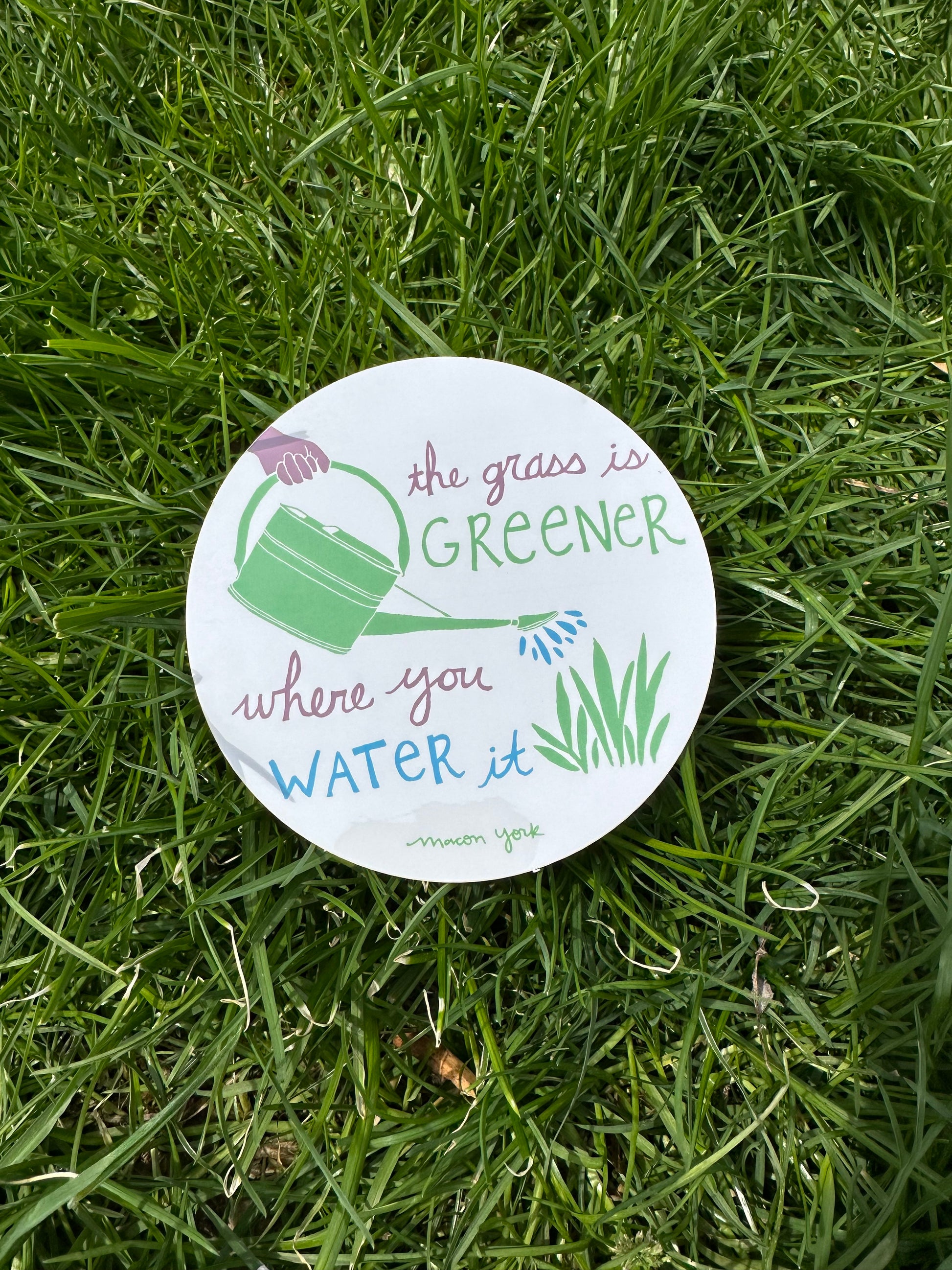 A 4" x 4" vinyl sticker featuring the words: The Grass is Greener Where You Water It" and a hand-drawn plum hand holding a green watering can, pouring blue water droplets on a patch of leafy grass. The text and original artwork hand-drawn by Macon York in her funky folk-art style. The sticker features three colors: a vibrant fern green, a rich plum, and a deep sky blue. Sticker is shown outside on the grass. 