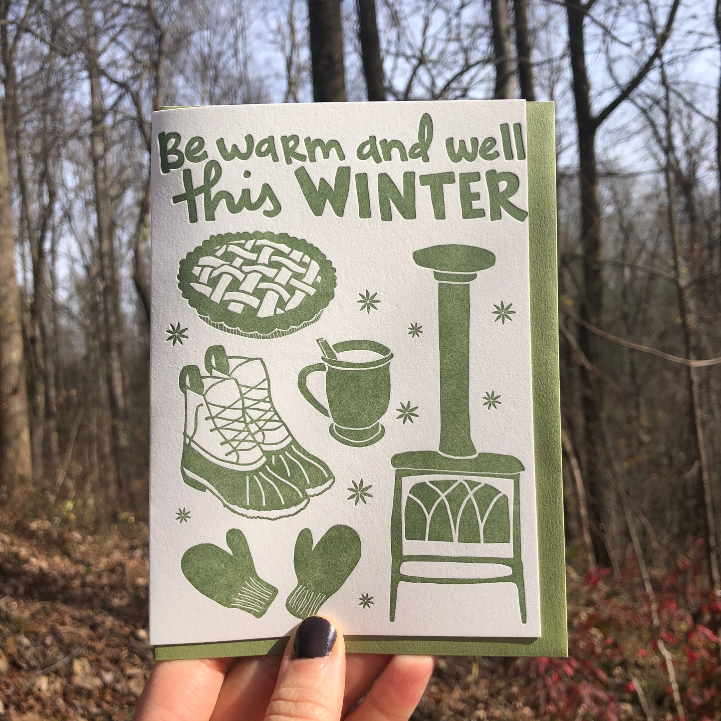 Celebrate Winter with this festive greeting card! This cheerful and cozy card features all the iconic image of the winter season: wood burning stove, star anise, hot cocoa (or cider!), fresh baked pie, mittens, winter boots. Hand-drawn images and typography are letterpress-printed in forest green ink. Green envelope.  Card is shown outside in front of a winter forest. 