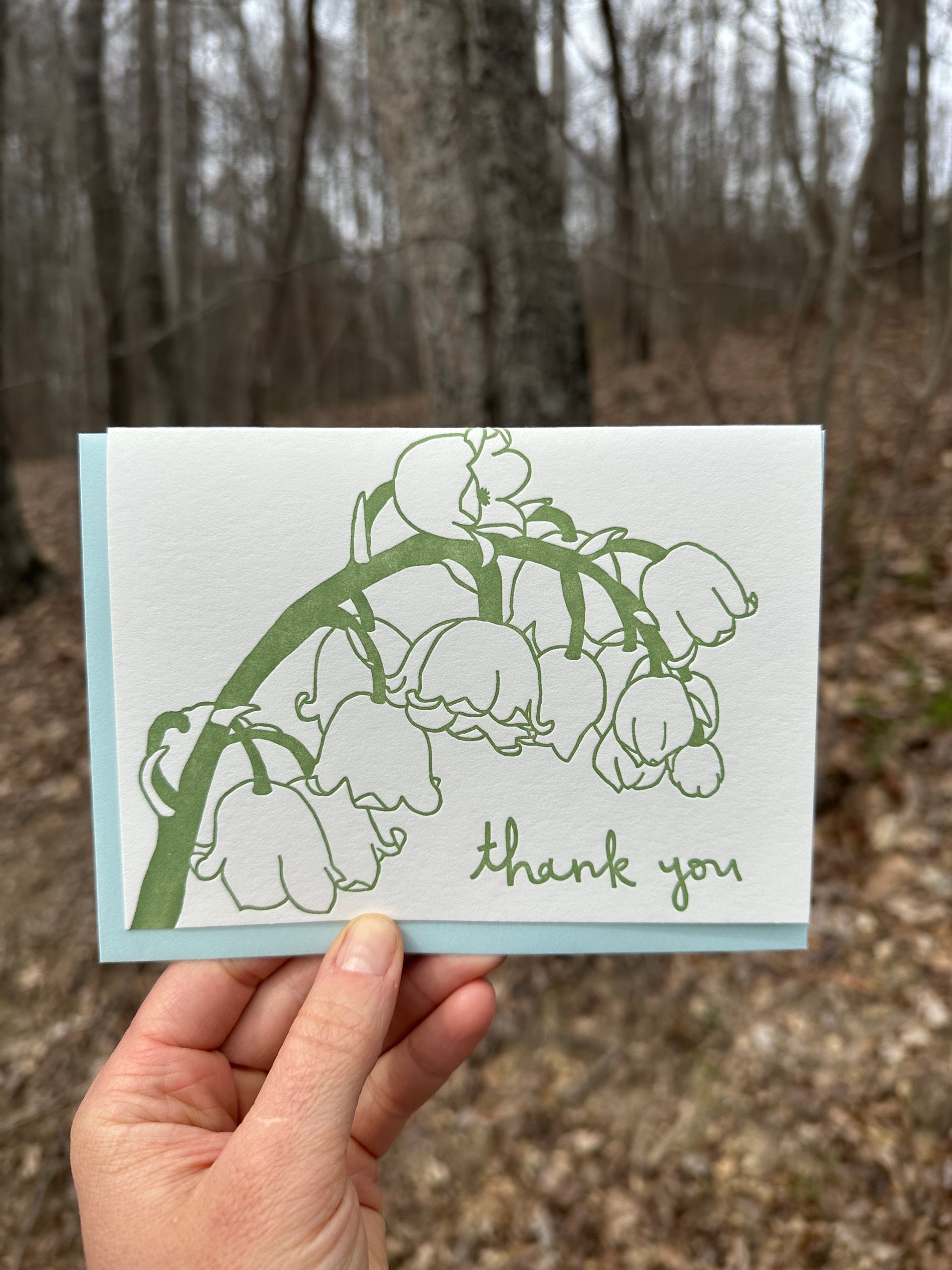 Letterpress greeting card featuring hand-drawn Lily of the Vlley, printed in a rich sage green ink. Whimsical hand-drawn text saying "Thank you" is shown at the bottom right side of the card, in the same green ink. The card is white, blank inside, and is paired with a light sky blue envelope. Card is shown outside in front of a deciduous forest in the winter. 