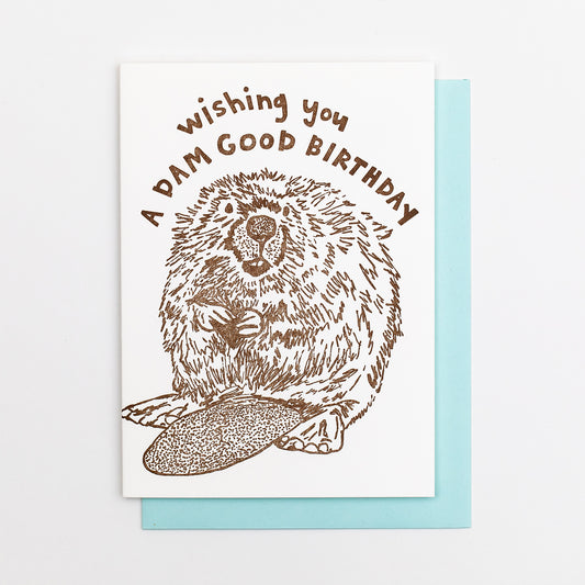 Letterpress greeting card featuring a hand-drawn Beaver printed in an earthy brown ink. "Wishing you a Dam Good Birthday!" is written on the top above the beaver in a whimsical hand-drawn text, in the same brown ink. The card is white, blank inside, and is paired with a light sky blue envelope.