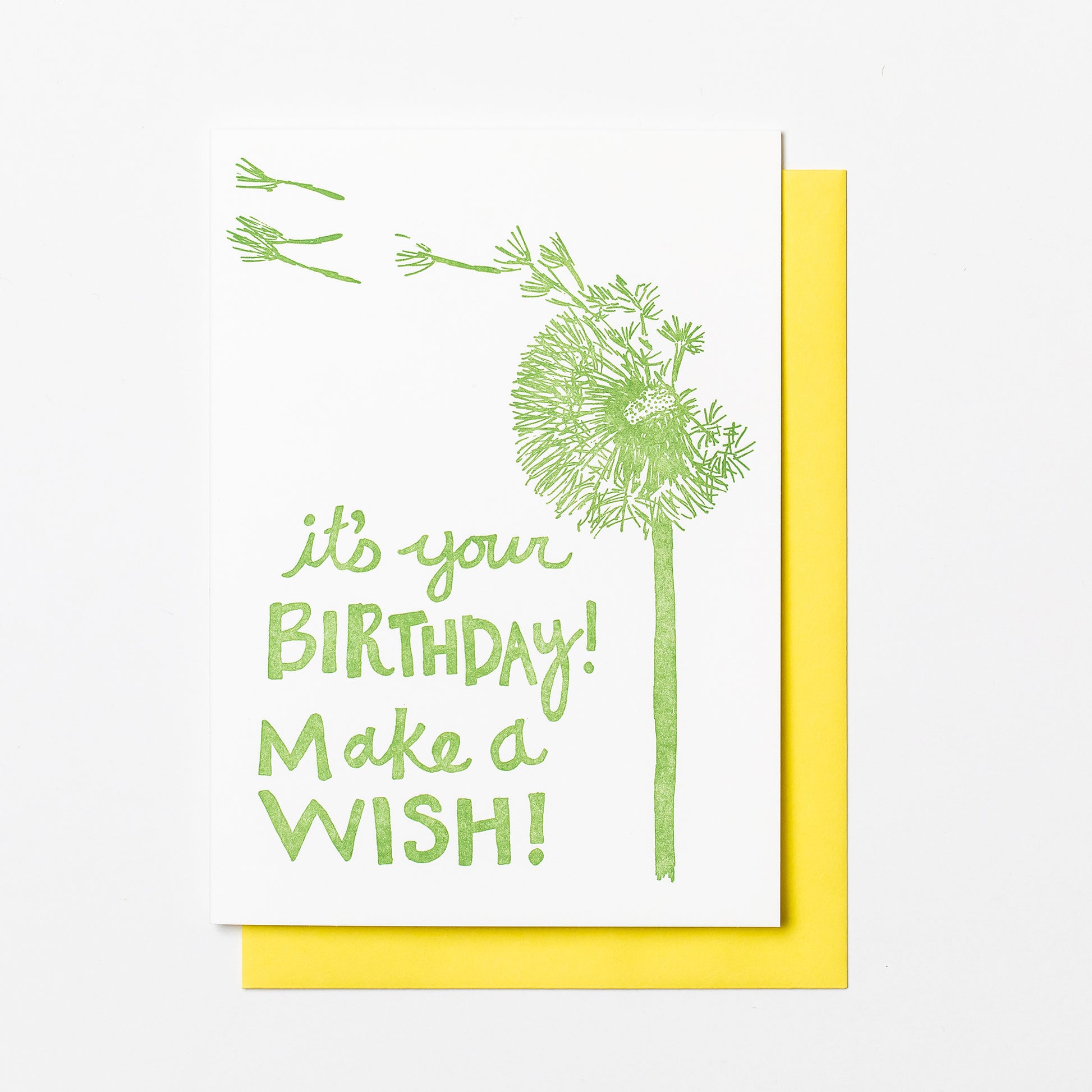 Letterpress greeting card featuring a hand-drawn dandelion printed in an earthy sage green ink. The bottom left of the card says "It's your birthday! Make a wish!" in a whimsical hand-drawn text, in the same earthy sage green ink. The card is white, blank inside, and is paired with a soft yellow envelope.