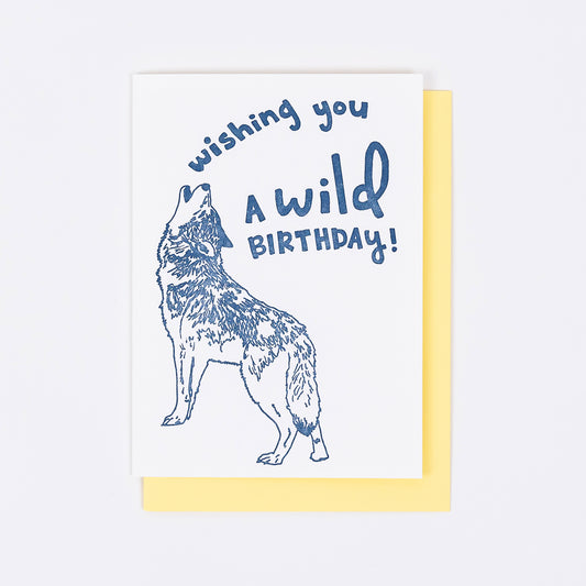 Letterpress greeting card featuring a hand-drawn Howling Grey Wolf printed in an earthy navy ink. "Wishing you a Wild Birthday!" is written on the top of the card in a whimsical hand-drawn text, in the same navy ink. The card is white, blank inside, and is paired with a soft yellow envelope.