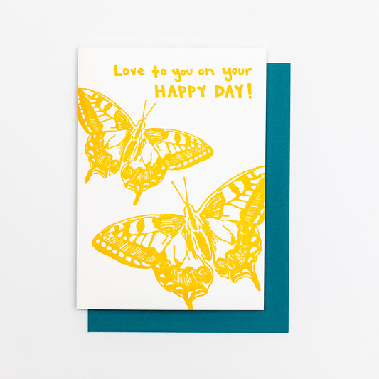 Letterpress greeting card featuring hand-drawn Eastern Swallowtail butterflies, printed in a vibrant golden yellow ink. Whimsical hand-drawn text saying "Love to you on Your Happy Day" is shown on the top of the card, in the same golden yellow ink. The card is white, blank inside, and is paired with a deep turquoise envelope.