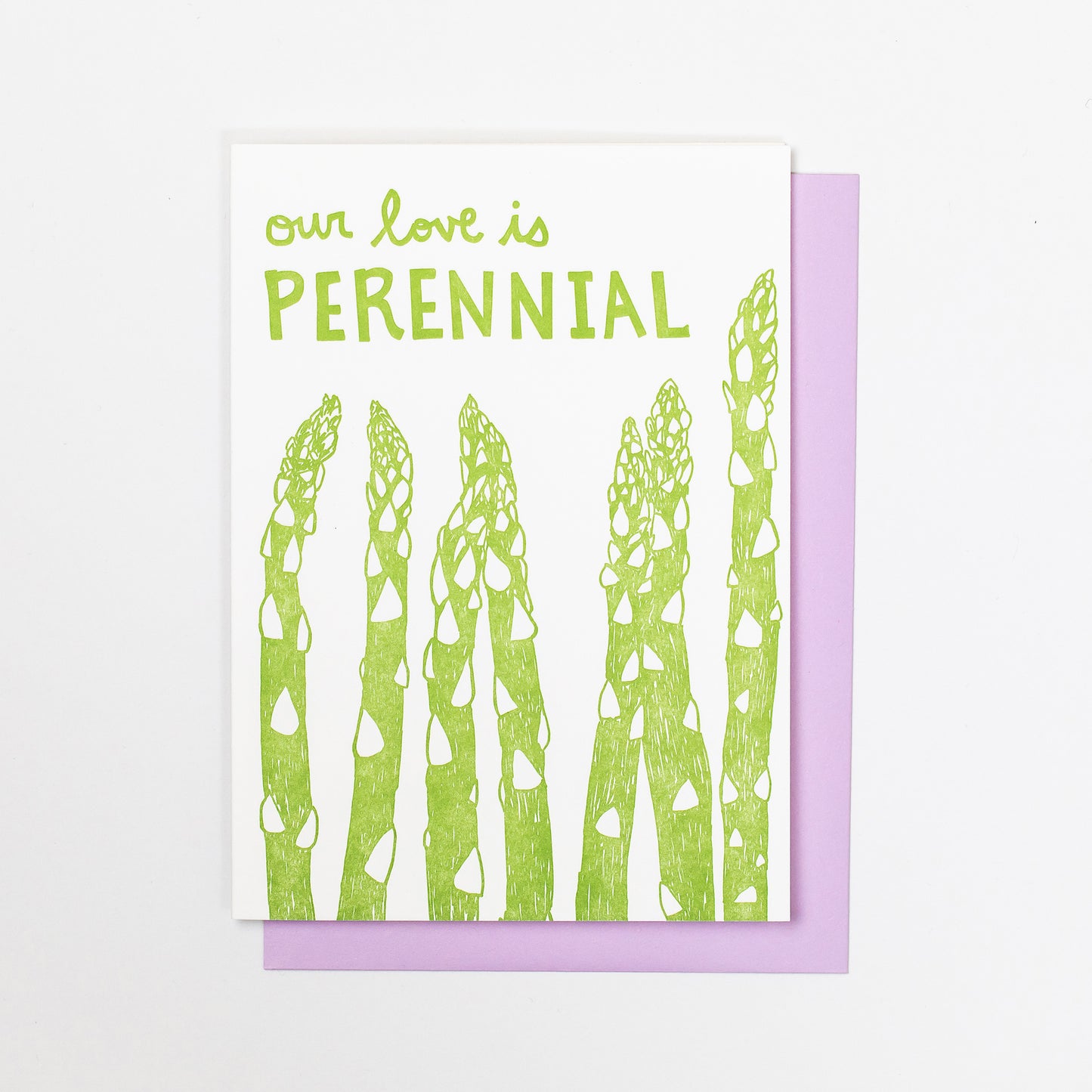 Letterpress greeting card featuring hand-drawn asparagus stalks, printed in a vibrant green ink. The top of the card says "Our love is perennial" in a whimsical hand-drawn text, in the same green ink. The card is white, blank inside, and is paired with a lilac purple envelope.