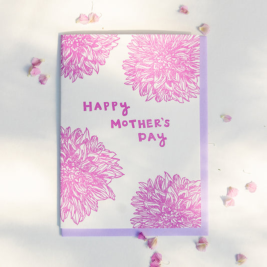 Letterpress greeting card featuring hand-drawn Dahlia flowers, printed in a vibrant dark fuchsia ink. The top center of the card says "Happy Mother's Day" in a whimsical hand-drawn text, in the same dark fuchsia ink. The card is white, blank inside, and is paired with a lilac purple envelope.