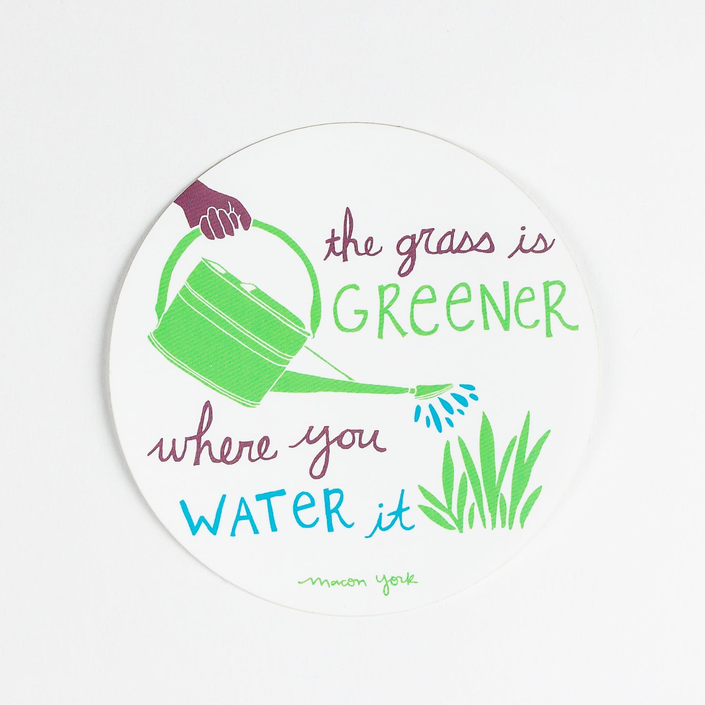 A 4" x 4" vinyl sticker featuring the words: The Grass is Greener Where You Water It" and a hand-drawn plum hand holding a green watering can, pouring blue water droplets on a patch of leafy grass. The text and original artwork hand-drawn by Macon York in her funky folk-art style. The sticker features three colors: a vibrant fern green, a rich plum, and a deep sky blue.