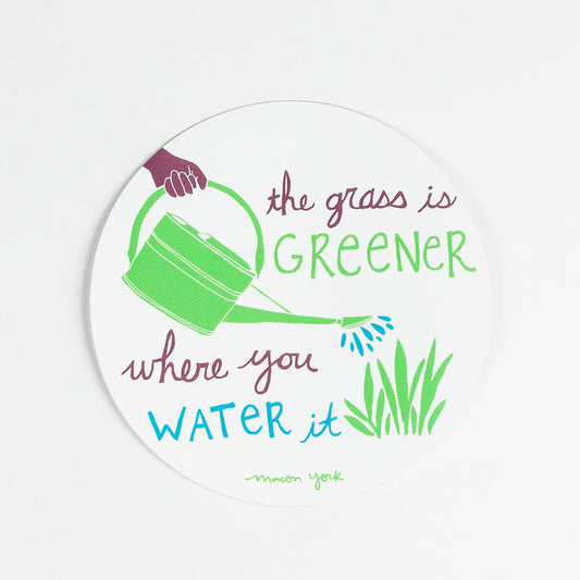 A 4" x 4" vinyl sticker featuring the words: The Grass is Greener Where You Water It" and a hand-drawn plum hand holding a green watering can, pouring blue water droplets on a patch of leafy grass. The text and original artwork hand-drawn by Macon York in her funky folk-art style. The sticker features three colors: a vibrant fern green, a rich plum, and a deep sky blue.