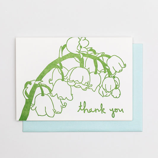 Letterpress greeting card featuring hand-drawn Lily of the Valley, printed in a rich sage green ink. Whimsical hand-drawn text saying "Thank you" is shown at the bottom right side of the card, in the same green ink. The card is white, blank inside, and is paired with a light sky blue envelope.