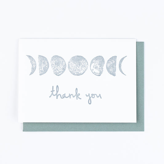 Letterpress greeting card featuring hand-drawn phases of the moon, printed in a shimmery silver ink. Whimsical hand-drawn text saying "Thank you" is shown centered on the card, in the same silver ink. The card is white, blank inside, and is paired with a slate gray envelope.