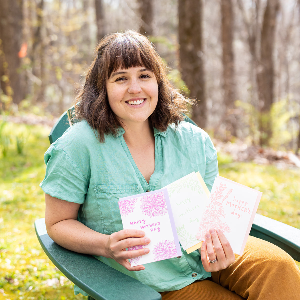 Macon York (adult female with short brown hair and bangs) sits outside in a green chair holding three Mother's Day Letterpress Greeting Cards: Dahlia, Brand, Wildflowers. She is smiling and looking directly at the camera. She is sitting in a grassy yard with deciduous trees behind her, in early Spring. 