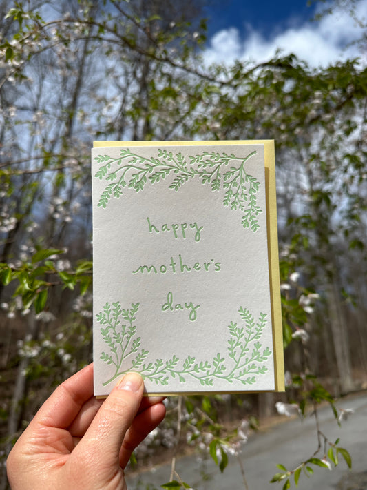 Letterpress greeting card featuring hand-drawn branches, printed in a soft minty green ink. The center of the card says "Happy Mother's Day" in a whimsical hand-drawn text, in the same minty green ink. The card is white, blank inside, and is paired with a warm yellow envelope. Card is shown outside on a spring day in front of a cherry tree.