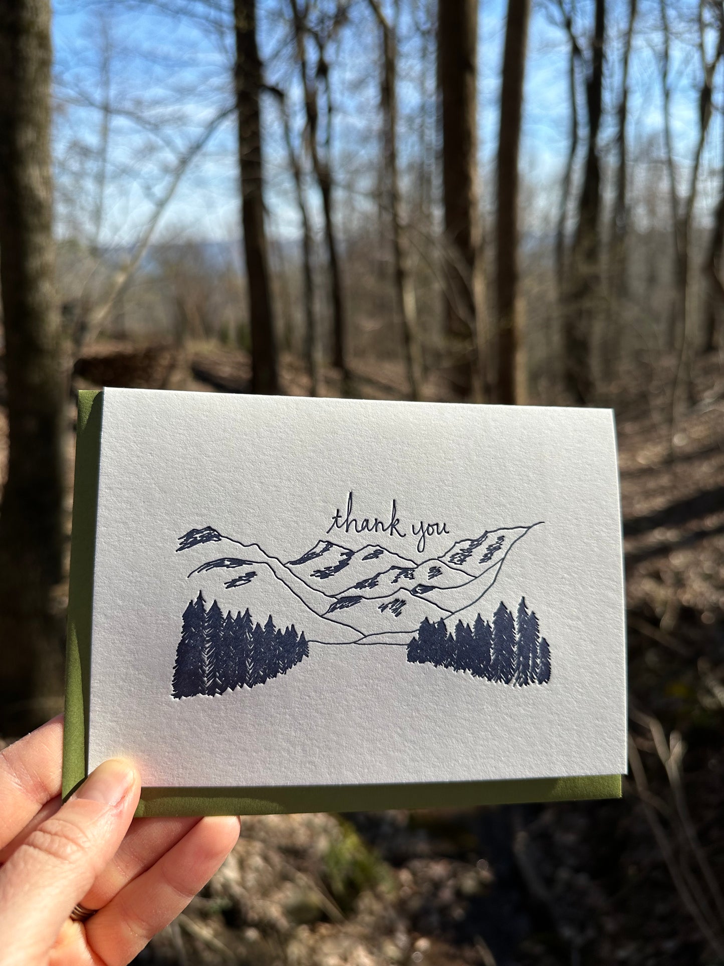 Letterpress greeting card featuring hand-drawn mountain landcape, printed in a rich navy ink. Whimsical hand-drawn text saying "Thank you" is shown above the moutains, in the same navy ink. The card is white, blank inside, and is paired with a rich green envelope. The card is shown outside on a sunny day in winter. 