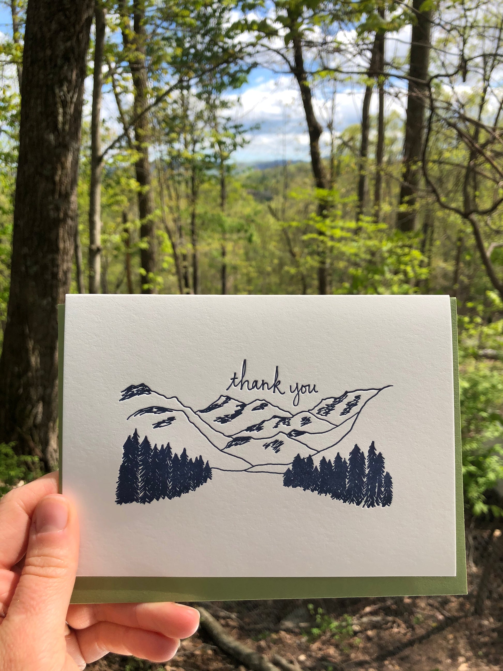 Letterpress greeting card featuring hand-drawn mountain landcape, printed in a rich navy ink. Whimsical hand-drawn text saying "Thank you" is shown above the moutains, in the same navy ink. The card is white, blank inside, and is paired with a rich green envelope. The card is shown outside on a sunny day in Spring. 