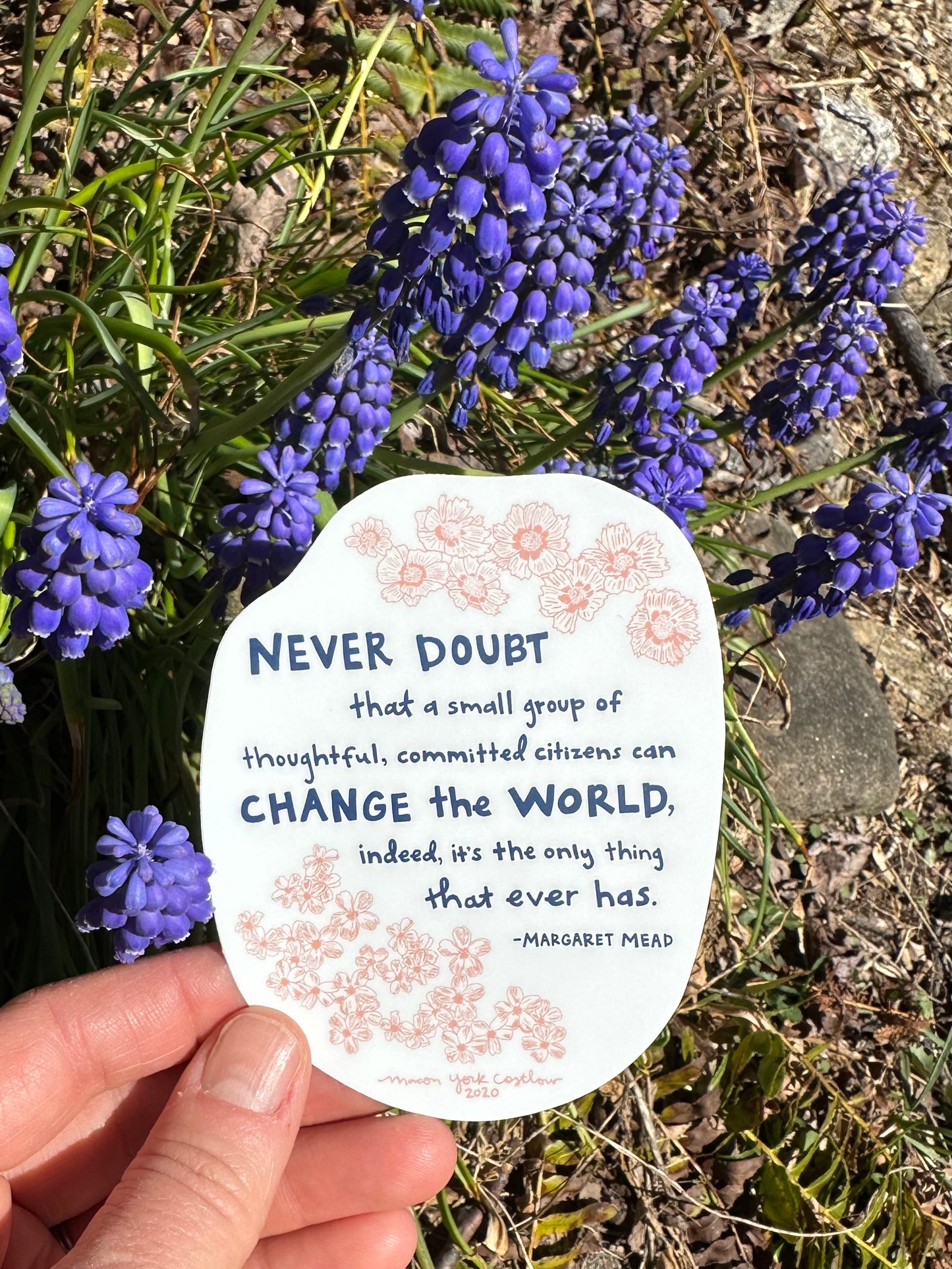 This vinyl sticker features original text by Margaret Mead, hand-drawn in a funky folk-art style of Macon York: "Never doubt that a small group of thoughtful, committed citizens can change the world, indeed, it's the only thing that ever has." Hand-drawn coreopsis and phlox in cheerful coral ink and hand-lettered text in navy.  Sticker is shown outside in front of blooming grape hyacinth. 