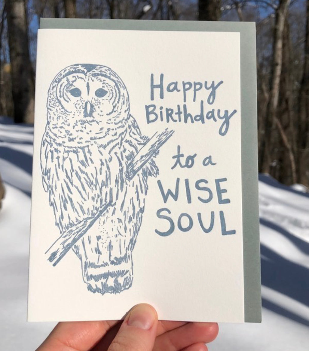 Letterpress greeting card featuring a hand-drawn barred owl printed in a lovely navy ink. The right side of the card says "Happy Birthday to a wise soul!" in a whimsical hand-drawn text, in the same navy ink. The card is white, blank inside, and is paired with a slate grey envelope. Card is shown outside in a snowy forest. 
