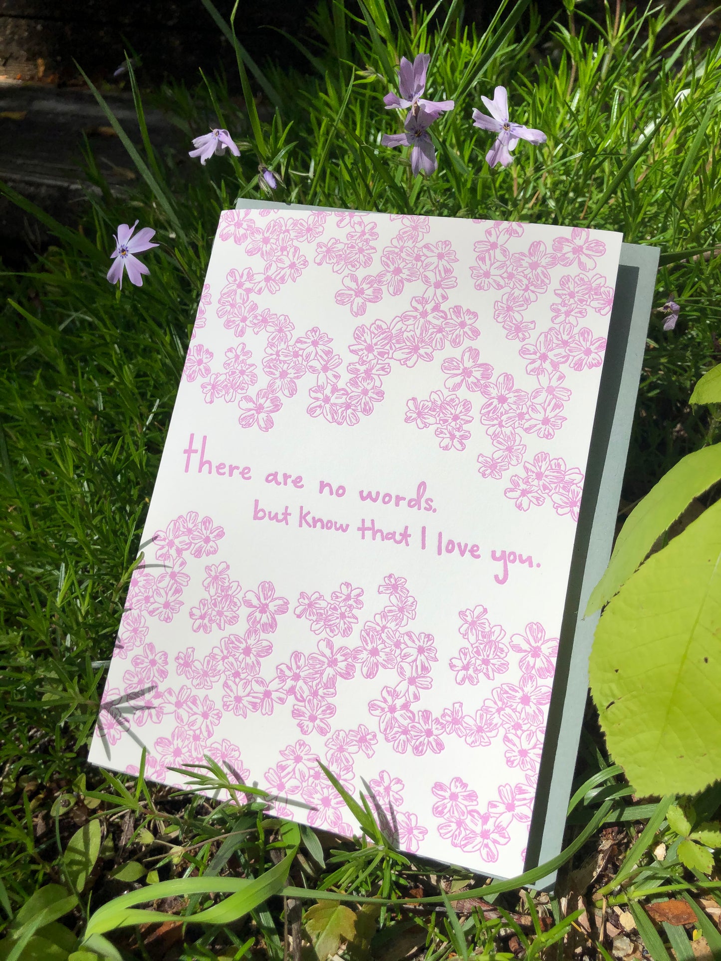 Letterpress greeting card featuring hand-drawn phlox flowers, printed in vibrant lavender ink. Whimsical hand-drawn text saying "There are no words, but know that I love you" is shown in the center of the card, in the same lavender ink. The card is white, blank inside, and is paired with a slate gray envelope. The card is shown outside with light purple phlox on a sunny spring day. 