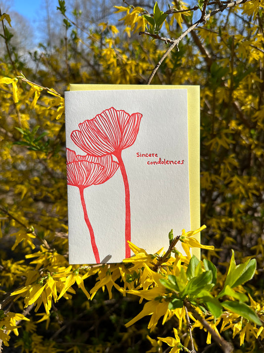 Letterpress greeting card featuring an intricate hand-drawn poppy flower, printed in a vibrant red ink ink. Whimsical hand-drawn text saying "Sincere Condolences" is shown on the right of the card, in the same red ink. The card is white, blank inside, and is paired with a soft yellow envelope. The card is shown in a blooming forsythia bush on a sunny spring day. 