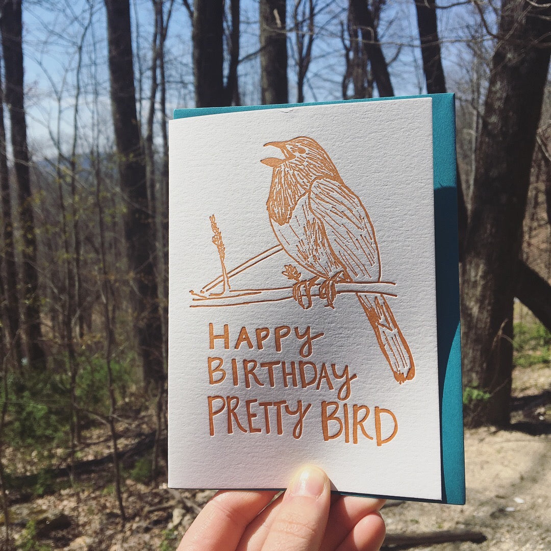 Letterpress greeting card featuring a hand-drawn Eastern towhee printed in a vibrant dark orange ink. The bottom of the card says "Happy Birthday Pretty Bird!" in a whimsical hand-drawn text, in the same dark orange ink. The card is white, blank inside, and is paired with a deep turquoise envelope. Card is shown outside in a forest on a sunny spring day.