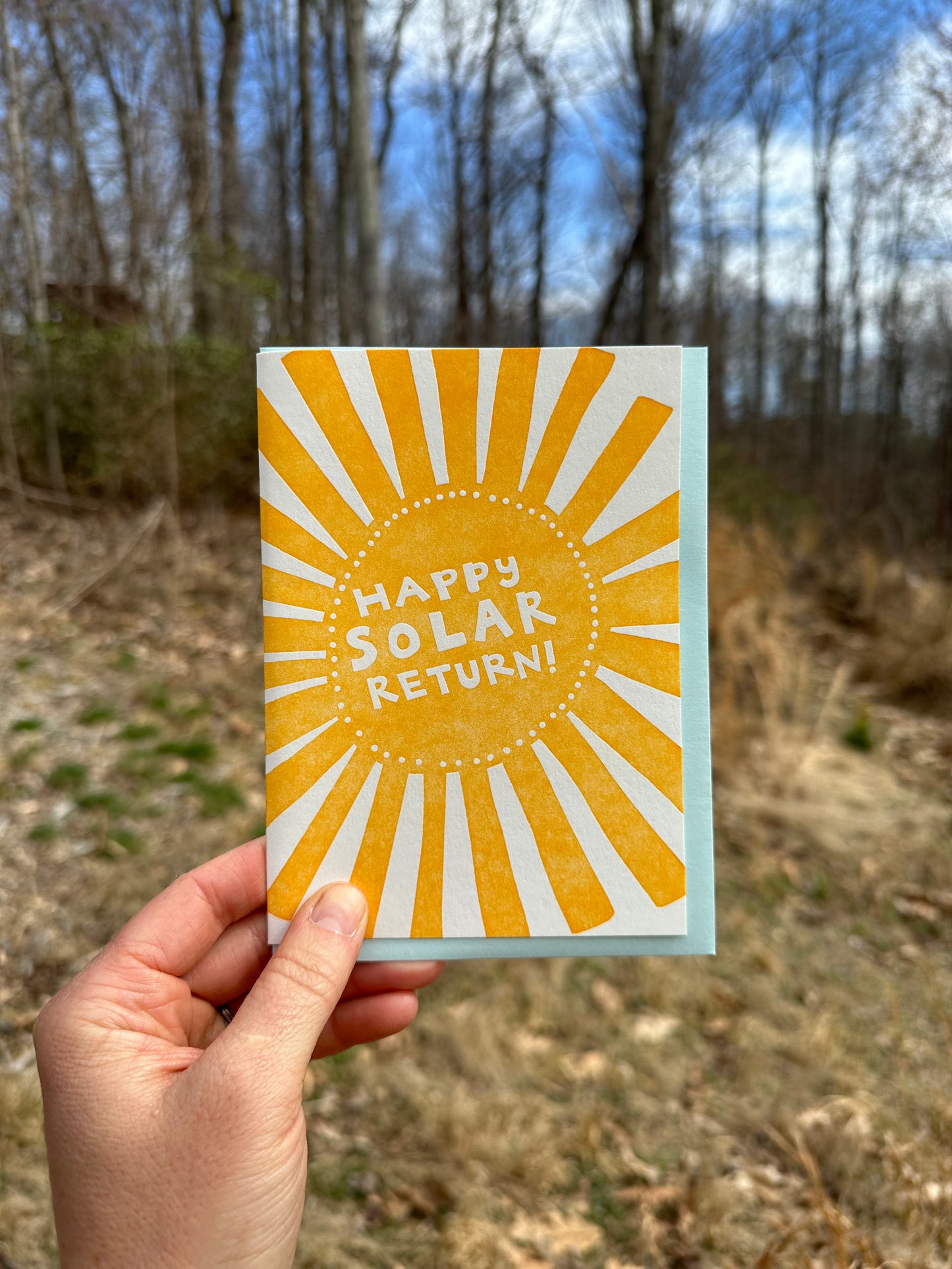 Letterpress greeting card featuring a hand-drawn Sun printed in a vibrant orange-gold ink. "Happy Solar return!" is written in the center of the sun in a whimsical hand-drawn text, in a knockout white. The card is white, blank inside, and is paired with a light sky blue envelope. Card is held by a hand in front of a field and forest on a sunny day. 