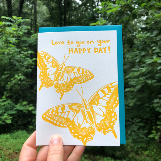 Letterpress greeting card featuring hand-drawn Eastern Swallowtail butterflies, printed in a vibrant golden yellow ink. Whimsical hand-drawn text saying "Love to you on Your Happy Day" is shown on the top of the card, in the same golden yellow ink. The card is white, blank inside, and is paired with a deep turquoise envelope. Card is shown outside in front of a lush forest in the summertime. 