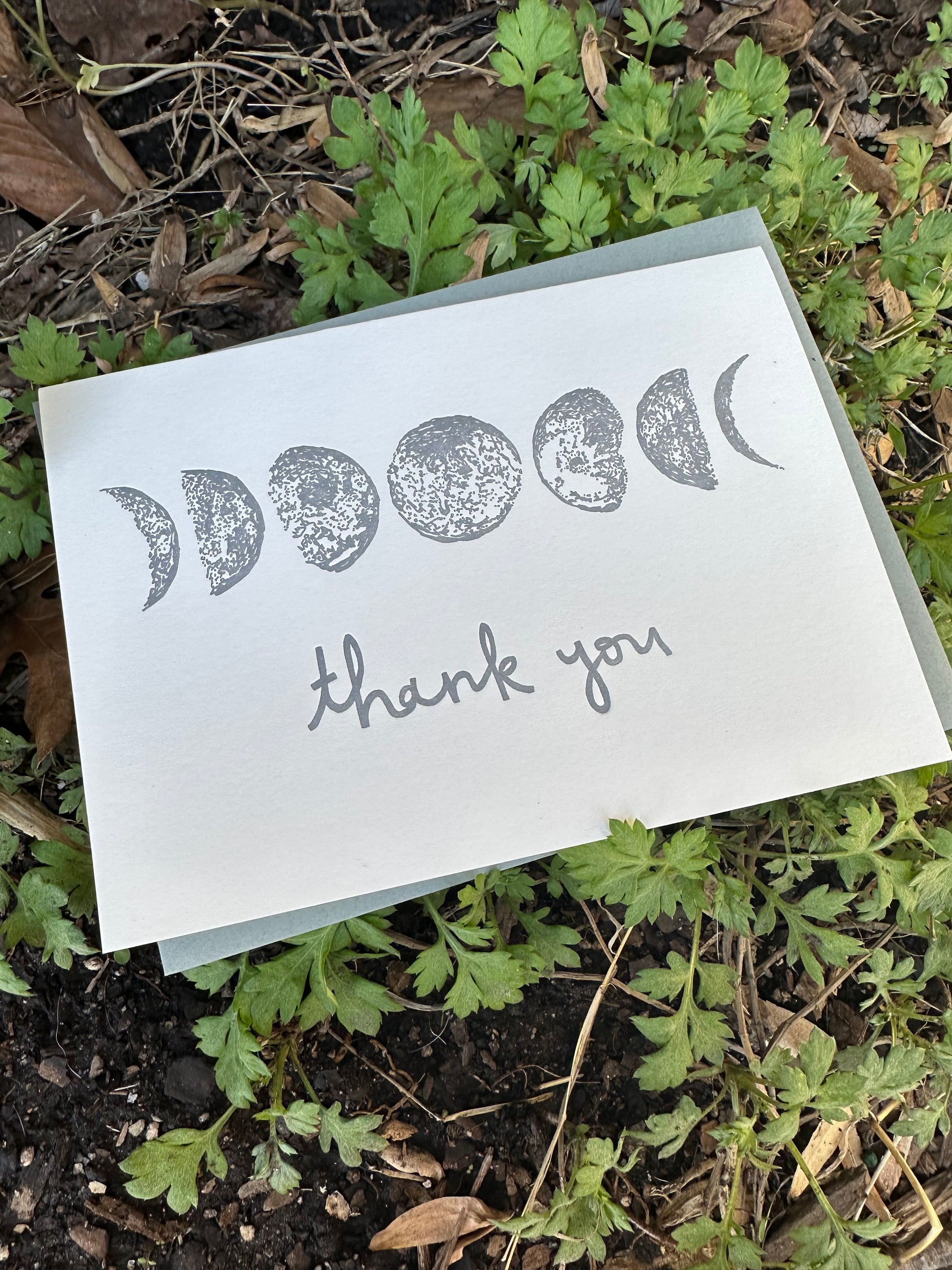 Letterpress greeting card featuring hand-drawn phases of the moon, printed in a shimmery silver ink. Whimsical hand-drawn text saying "Thank you" is shown centered on the card, in the same silver ink. The card is white, blank inside, and is paired with a slate gray envelope. The card is shown outside on a bed of mugwort.