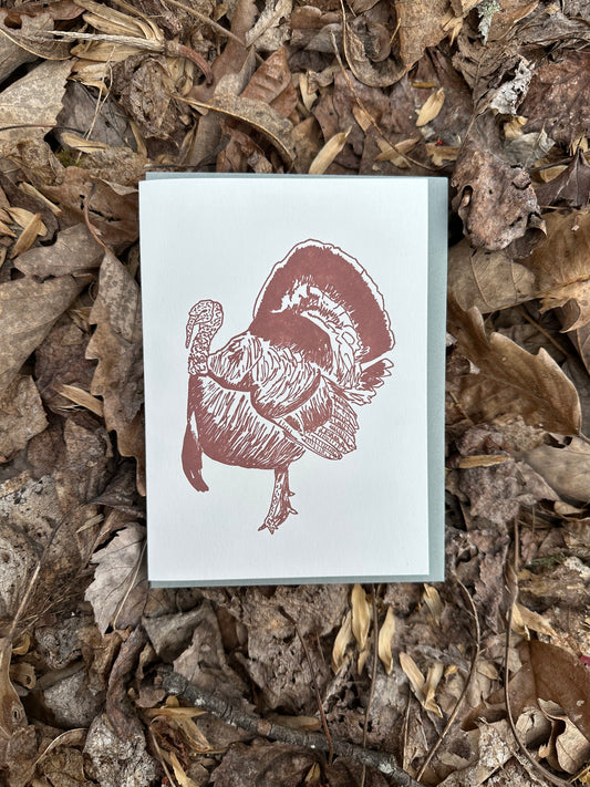 Letterpress greeting card featuring hand-drawn Appalachian Wild Tom Turkey, printed in an earthy brown ink. There is no text on the card. The white card is 100% cotton, blank inside, and is paired with a slate gray envelope. The card is shown outside on a bed of fall leaves. 