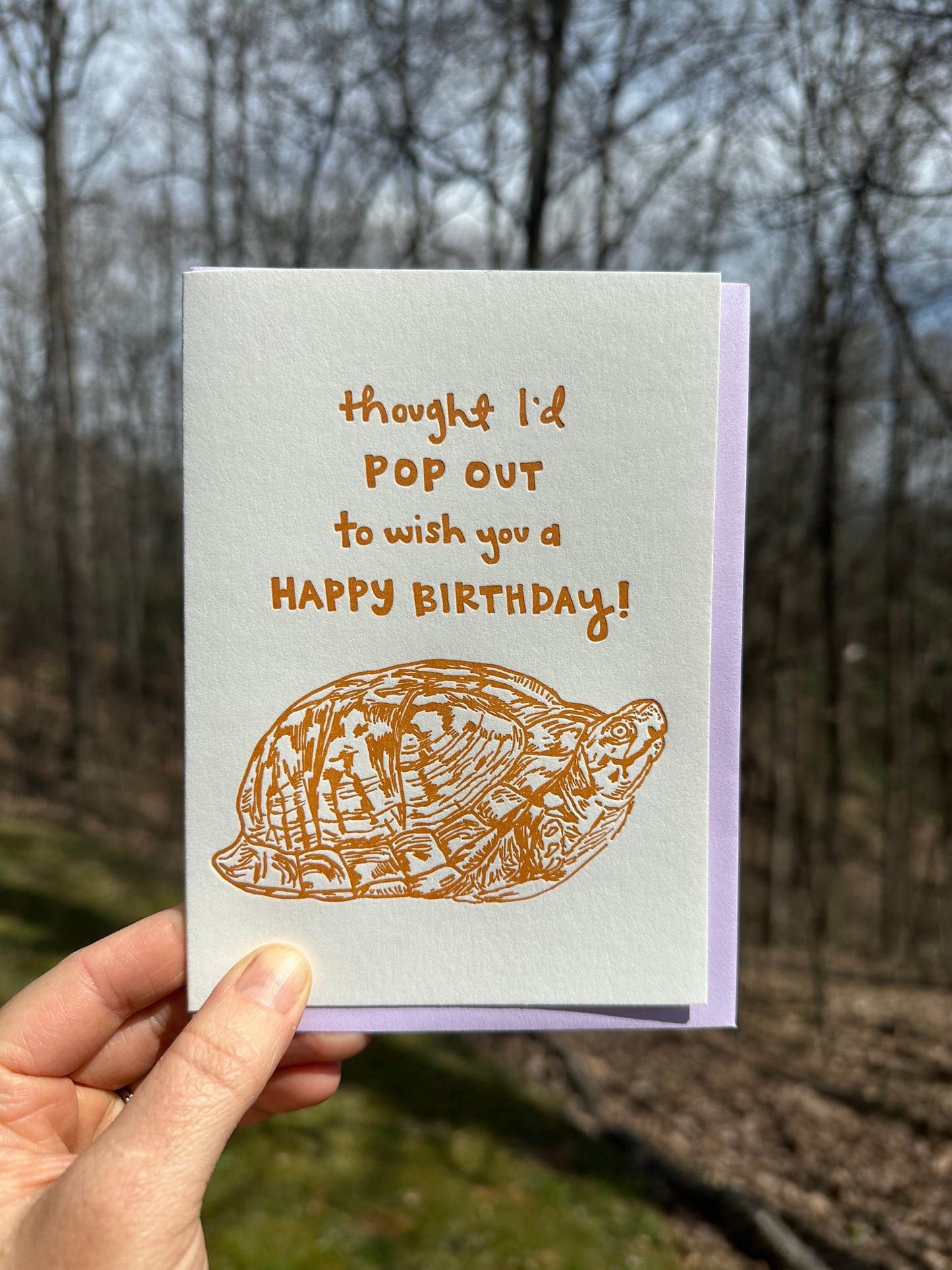 Letterpress greeting card featuring a hand-drawn Appalachian Box Turtle printed in a vibrant dark orange ink. "Thought I'd pop out to wish you a Happy Birthday!" is written on the top of the card in a whimsical hand-drawn text, in the same orange ink. The card is white, blank inside, and is paired with a lilac purple envelope. Card is held up outside in front of a spring forest on a sunny day. 