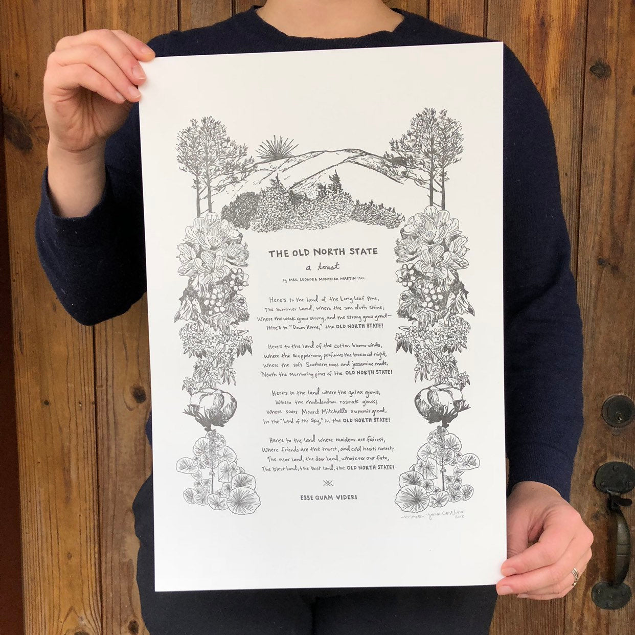 Large letterpress art print featuring "The Old North State," an ode to North Carolina and all her natural beauty, written in 1904 by Mrs. Leonora Monteiro. Macon York has hand-written the entire toast in her whimsical style. All the native plants in the toast are hand-drawn to create a border around the text.  The artist, Macon, is shown holding the print - a zoomed in shot of just the print and her hands. 