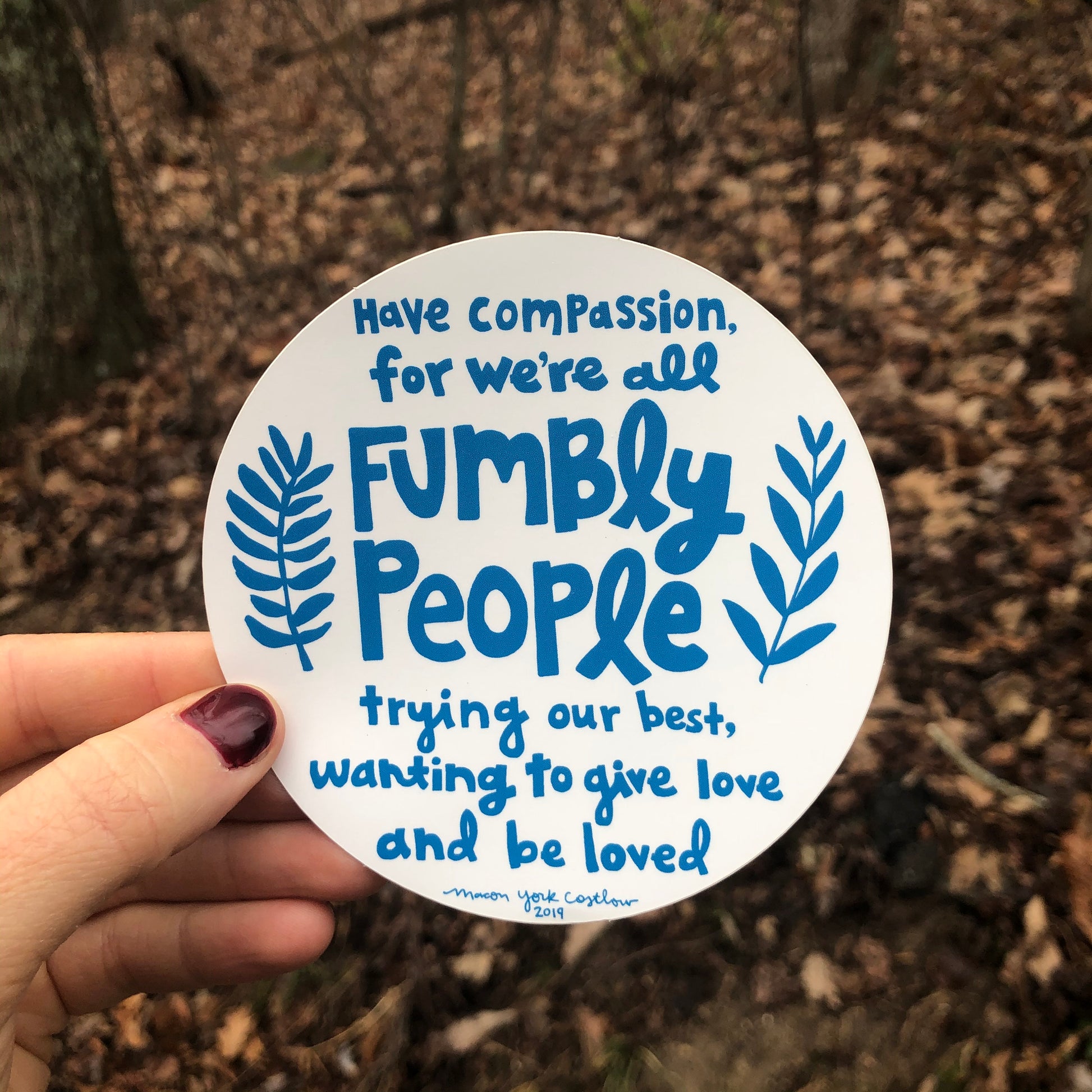 This vinyl sticker features original text by Macon York, hand-drawn in a funky folk-art style. 4" x 4" Circle Vinyl Sticker.  Text reads "Have compassion, for we're all fumbly people trying our best, wanting to give love and be loved." Sticker is shown outside in front of a winter forest. 